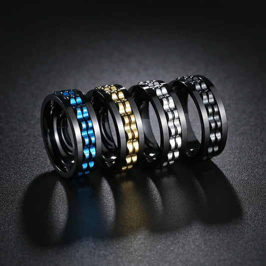 Four metallic rings in black, gold, silver, and blue on a dark textured surface