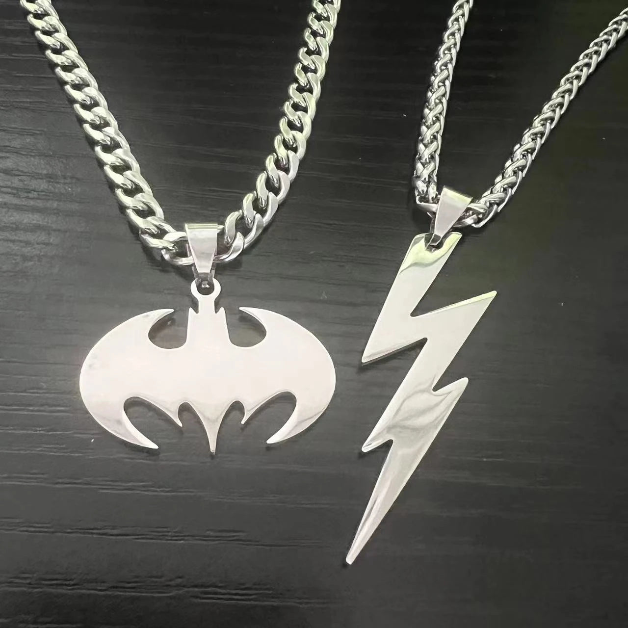 Two silver pendants, one resembling a bat symbol and the other a lightning bolt, on a dark wooden surface.